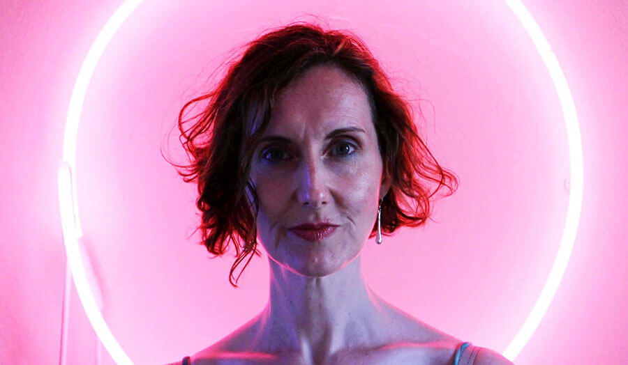 Suzanne Cohen, a female lecturer, stands against a pink background looking at the camera.