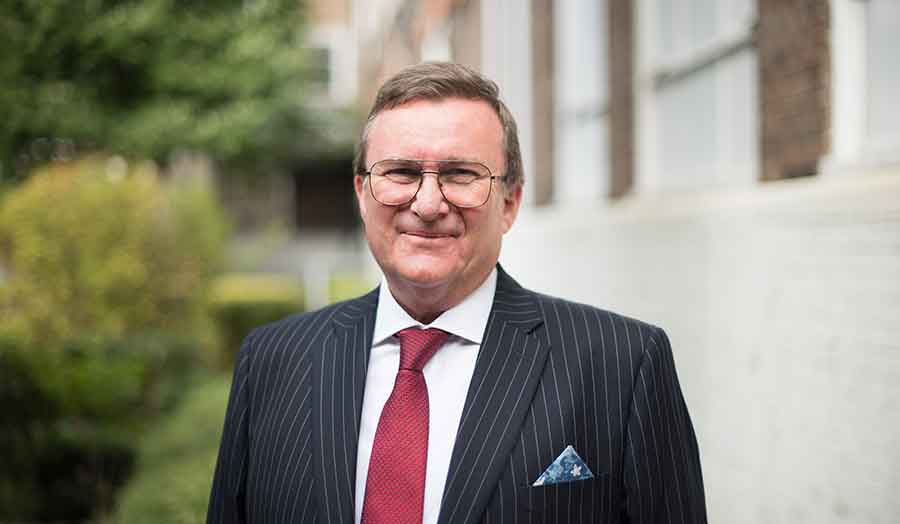 A faculty member wearing suit standing in front of university buildings.