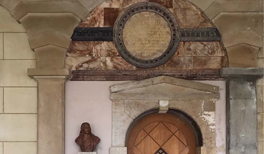 Stone arch with ornate circular plaque set in marble over wooden door and bust of a man on pedestal