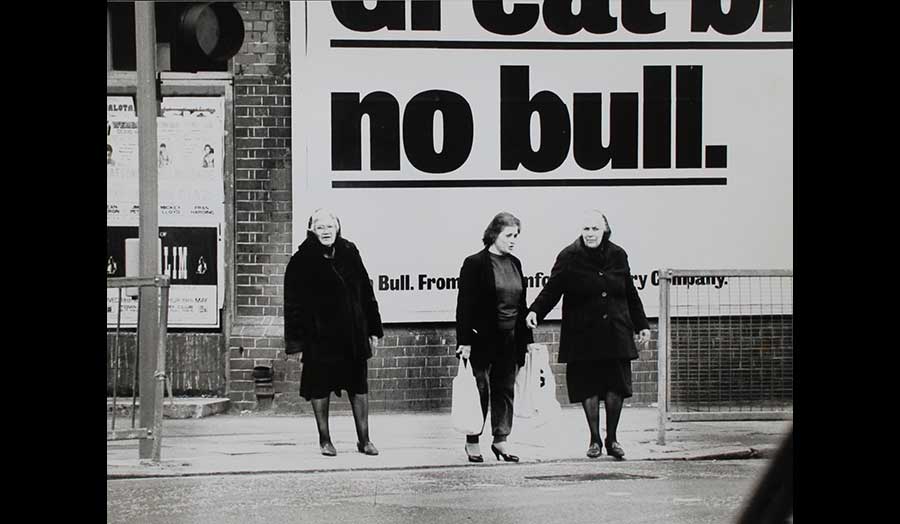 Three women stand on pavement under a large advertisement.