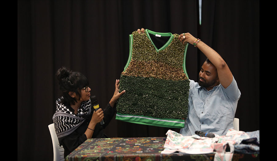 Two people in discussion hold up a green and highly textured garment in front of an audience