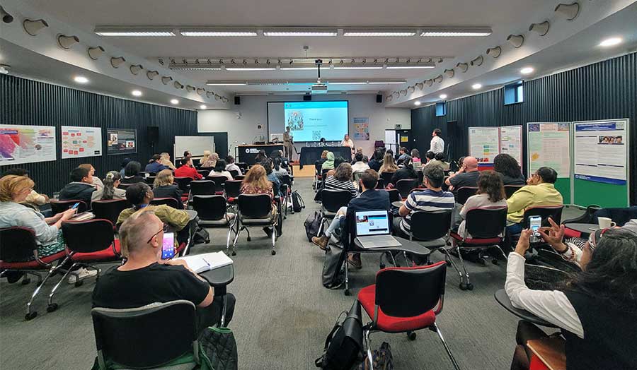 A room full of students and staff watching a research presentation, with research posters on the walls