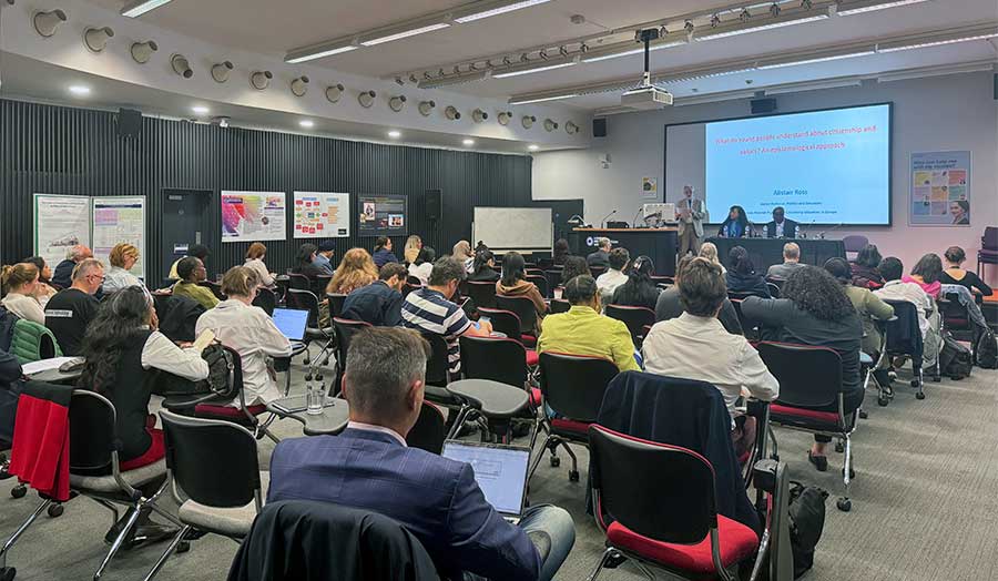 A room full of students and staff watching a research presentation, with research posters on the walls