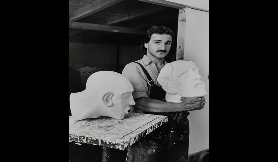 A man in overalls stands in a studio holding a large model of a human head
