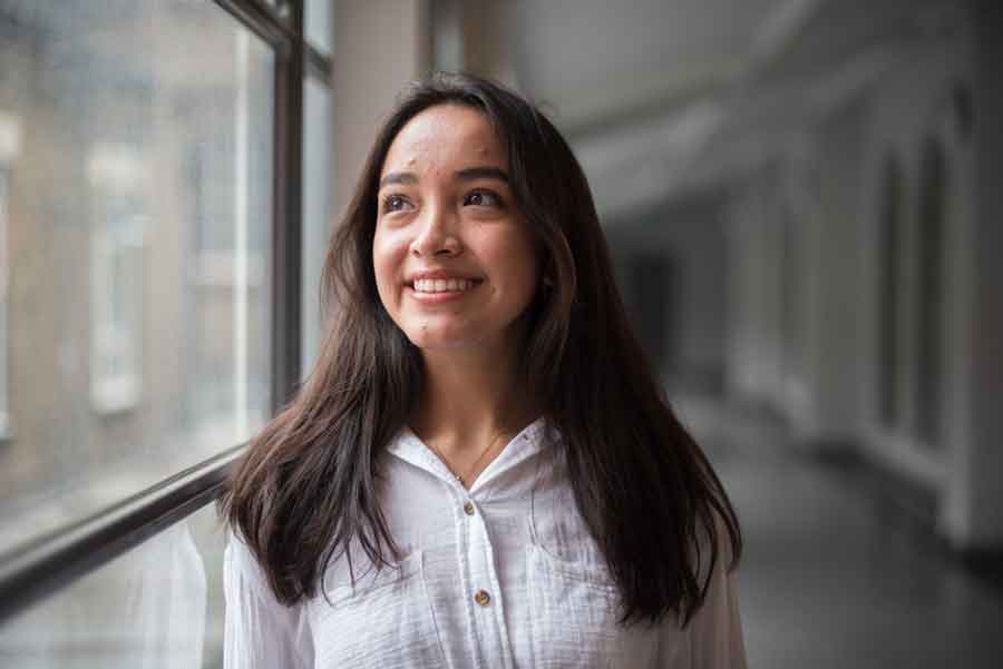 A smiling student standing next to a window in a corridor